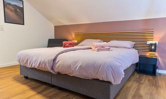 A cozy bedroom with a large bed and soft, pink bed sheets. Next to the bed is a nightstand with a lamp.