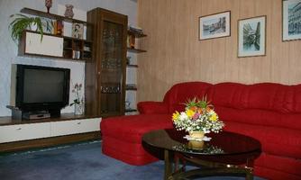 A cozy living room with a red sofa and a round table. Pictures are hanging on the wall, and in the background, there is a television.