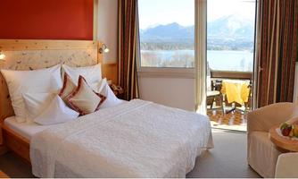 A cozy bedroom with a large bed and colorful pillows. The view from the window shows mountains and a lake.