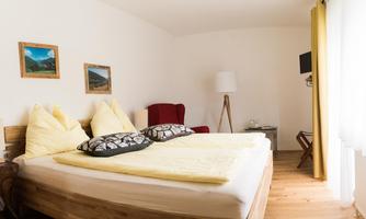 A cozy bedroom with a large bed, yellow pillows, and wooden flooring. The room features a seating area and some pictures on the wall.