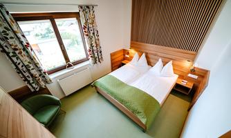 A modern hotel room with two beds and wooden paneling. Large windows provide plenty of natural light and a view outside.