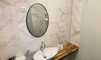 A modern bathroom with a round mirror and a stylish sink. The walls are clad in elegant marble tiles, and a wooden shelf complements the design.