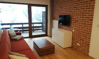 A modern living room with a red sofa and a wooden table. Large windows offer a view of a balcony and showcase a cozy space with a stone wall.