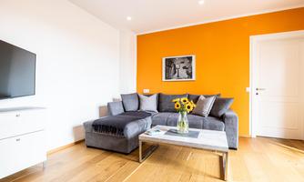 A modern living room with a gray sofa and an orange accent wall. On the table, there is a vase with sunflowers, and there is a TV on the wall.