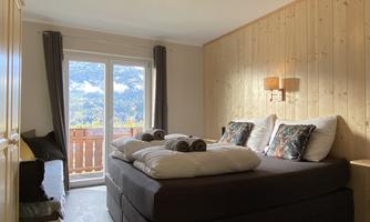 A modern bedroom with a large double bed and natural wood. Large windows provide a view of the surrounding mountains.