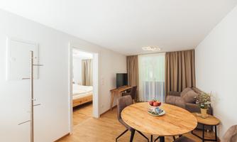 A bright living area with a round wooden table and comfortable chairs. In the background, there is a television and access to a bedroom with curtains.