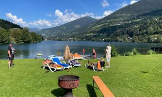 A picturesque lake landscape with green meadows and mountains in the background. People relax on loungers and enjoy the sun by the water.