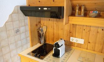 A modern kitchen with wooden shelves and a small cooking area. An espresso machine stands on the countertop, and kitchen utensils are stored in a container.