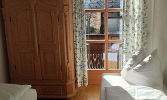 A cozy bedroom with two beds and a large wooden wardrobe. A lot of daylight is visible through the window.