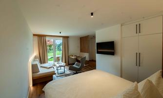 A modern hotel room with a cozy double bed and a small seating area. Large windows offer a view of the outdoor area and plenty of natural light.