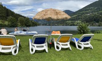 A beautiful meadow by the water with loungers and a sun umbrella. In the background, mountains and a boat can be seen.