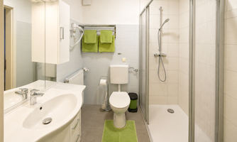 A modern bathroom featuring a shower, a sink, and a toilet. The walls are brightly tiled, and green towels along with a rug give the space a fresh accent.