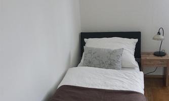 A simple bedroom with a single bed and white bedding. Two pictures hang on the wall, and next to the bed is a bedside lamp.