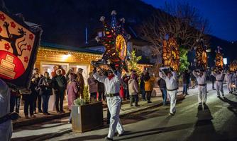 Eine festliche Parade bei Nacht mit Menschen in traditionellen Kostümen, die große Lichterträger tragen. Zuschauer stehen am Straßenrand und beobachten das Ereignis.
