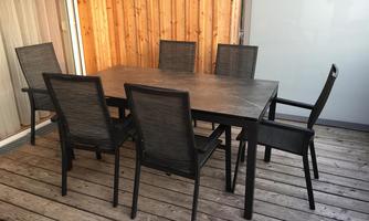 A large table with six chairs stands on a wooden terrace. The wall in the background is made of wood and the space feels bright and inviting.