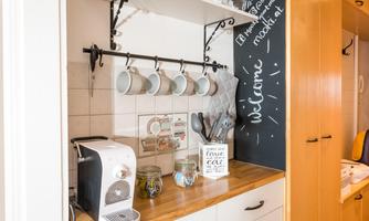 A modern kitchen unit with a wooden countertop and a coffee machine. Cups hang on the wall, and there are decorative elements on the table.