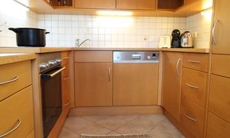 A modern kitchen with wooden cabinets and a bright tiled floor. Cooking utensils and electrical accessories are on the countertop.