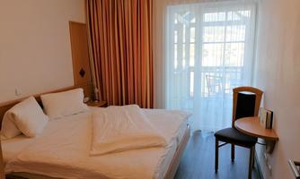 A bright bedroom with a large bed and a view of a balcony. There is a small table and a chair next to the window.