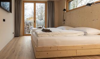 A modern bedroom with a large double bed and wooden furniture. The large windows provide plenty of daylight and offer a view of the nature outside.
