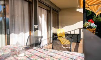 A cozy balcony with a colorful table and a yellow lounge chair. The sun is shining on the white curtains and the flowers are lively in a pot.