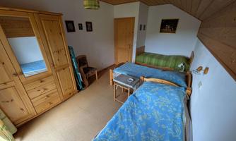 A cozy bedroom with two beds and a wooden wardrobe. The walls are simply designed and there is a window that provides natural light.