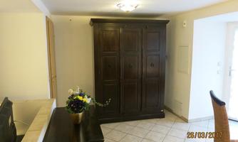 A stylish living space with a large, dark wardrobe and a simple table. The floor is tiled and there is plenty of natural light.