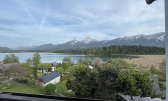 A picturesque view of a lake surrounded by mountains. In the foreground, green meadows and several buildings can be seen.