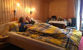 A cozy bedroom with a double bed and wooden paneling. Two people are lying under a colorful blanket and seem to be happy together.