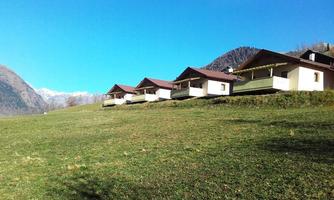 A group of huts stands on a green meadow with a view of the mountains. The sky is clear and blue, creating a tranquil atmosphere.