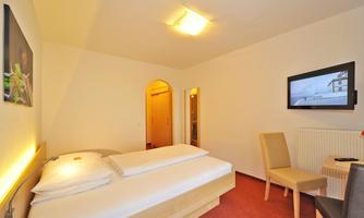 A simple and modern bedroom with a double bed, a television, and two chairs. The walls are light, and the floor is carpeted.