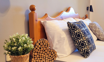 A cozy bedroom with a bright wooden bed and decorative cushions. A decorative plant pot and a heart made of wooden logs are on the nightstand.