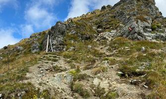 A steep hiking trail leads through rocky terrain. Above the path, the sky is partly cloudy and blue.