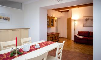A cozy dining area with a white table, chairs, and a red tablecloth. In the background, there is a living room with a red sofa and warm light.