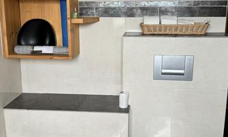 A bathroom with a white bathtub and a modern toilet. On the wall are wooden shelves with various bottles and a basket.