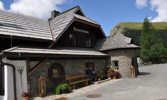 A traditional house with a stone facade and a sloping roof. There are flowers and plants in front of the house, as well as a beautiful mountain landscape in the background.
