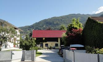 A charming house with a red roof and surrounding trees. In the background, green mountains and blue sky can be seen.