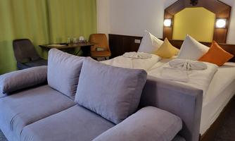 A cozy hotel room with a double bed and a gray sofa. The walls are in neutral tones, and a green curtain adds a touch of fresh color.