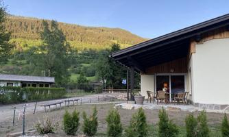 A modern house with a large garden in a mountainous landscape. In the foreground, small shrubs and a seating area on the terrace are visible.