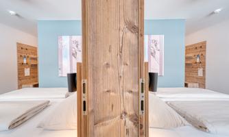 A modern bedroom with two single beds and a wooden partition wall. The walls are in soft color tones, and there are decorative elements in the background.