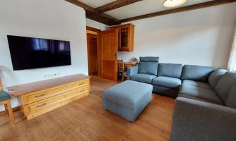 A bright living room with a gray sofa and a wooden TV table. There is a large television on the wall and a cozy armchair in the corner.