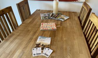 A wooden table with several chairs is depicted. On the table lie two informational brochures, a notebook, and some postcards, as well as a large candle in the center.