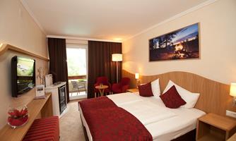 A cozy hotel room with a large bed and red accents. There is a television, a table and armchair next to the bed, as well as a balcony with a view outside.