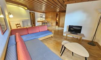 A cozy living room with a wooden ceiling, a red and blue sofa, and a television. The room is bright and welcoming, with a table and chairs in the background.