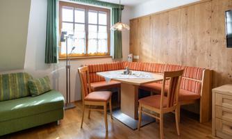 A cozy dining area with wooden furniture and a round table. The seating area is equipped with orange cushions and provides plenty of light through the large windows.