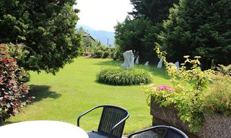 A beautiful garden with green grass, sculptures, and colorful plants. In the foreground, there are chairs and a table on a terrace.