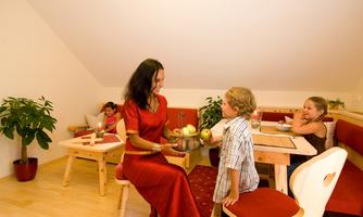 A woman is sitting at a table with a boy and is handing him apples. In the background, two children are playing while plants decorate the room.