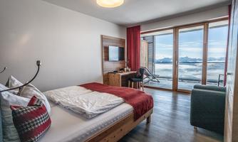 A modern hotel room with a large window and a view of the mountains. It is cozily furnished with a bed, a table, and an armchair.