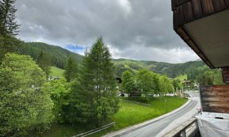 A view of a green landscape with trees and hills under a cloudy sky. The road runs along the meadow.