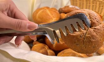 A hand holds a pair of tongs to take a roll from a basket. In the background, many freshly baked rolls are visible.
