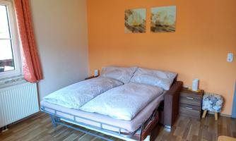 A cozy bedroom with a large bed and an orange wall. Next to the bed is a bedside table and a chair, and there are windows with curtains.